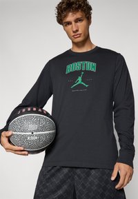 Sort langærmet skjorte med grøn tekst "BOSTON", der har et basketball-grafik. Grå basketball med tekstureret overflade og røde detaljer holdt i hånden.