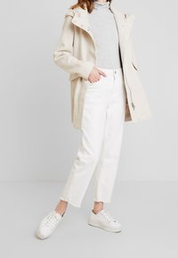 Beige stickad kappa med huva, grå polotröja, vita jeans med råa kanter och vita sneakers. Kappan har tryckknappar och en avslappnad passform.