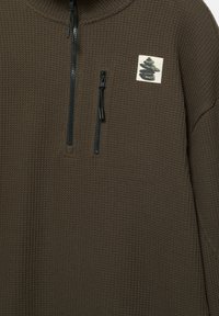 Hnedý pulóver na zips s textúrovaným vzorom pripomínajúcim wafle, s malou grafickou nášivkou a čiernym zipsom.