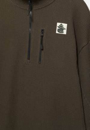 Maglione marrone con cerniera frontale, caratterizzato da un design strutturato a nido d'ape, con una piccola toppa grafica e dettagli della cerniera nera.