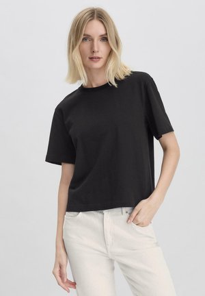 Schwarzes Kurzarm-T-Shirt in lockerer Passform, aus weicher Baumwolle. Mit einem Rundhalsausschnitt und minimalen Nähdetails, kombiniert mit hellen Hosen.