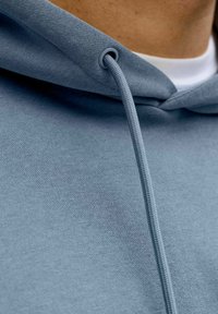 Svetlo modra jopa s kapuco z mehko teksturo, kenguru žepom in vrvico skozi siv obroček na ovratniku.