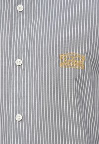 Siva srajca z belimi navpičnimi črtami iz blaga. Ima bele gumbe in zlat vezen logotip.