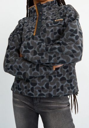 Person iført en grå og sort mønstret fleece-pullover med knapper foran og Columbia-logo, kombineret med mørkegrå jeans.