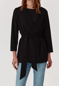 Svart omslagjacka i mjuk tyg, med långa ärmar, ett knytbälte i midjan och en avslappnad passform. Matchad med ljusblå jeans.