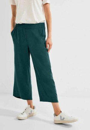 Personne portant un pantalon court large vert foncé avec une ceinture élastique et des baskets blanches avec des accents gris.