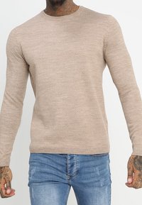 Beżowy dziergany sweter z okrągłym dekoltem i długimi rękawami, z teksturowanym wzorem i ściągaczem u dołu, w zestawie z jasnoniebieskimi dżinsami.