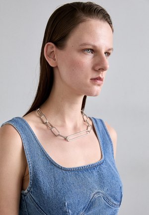 Kobieta z prostymi brązowymi włosami, ubrana w bezrękawnik z denimu i duży srebrny łańcuch, zwrócona w prawo na jednolitym tle.