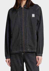 Muž oblečený v černé pruhované zipové bundě a odpovídajících kalhotách s viditelným logem adidas Trefoil na hrudní nášivce.