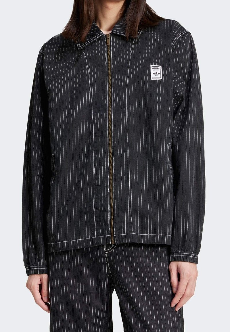 Muž oblečený v černé pruhované zipové bundě a odpovídajících kalhotách s viditelným logem adidas Trefoil na hrudní nášivce.