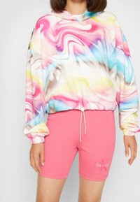 Multicolore hoodie met spiraalpatronen in roze, blauw en geel, voorzien van een trekkoord. Gecombineerd met roze katoenen shorts met tekst.