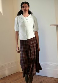 Cardigan tricoté vert clair avec un col en forme de vague et des boutons, associé à une longue jupe en tartan marron avec des plis et une fente.