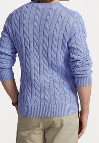 Person som bär en ljuslila kabelstickad tröja med uppvikta ärmar och beige byxor, sedd bakifrån mot en enfärgad bakgrund.