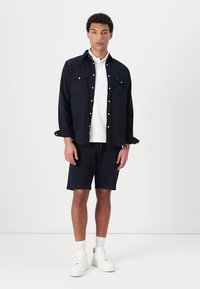 Cămașă albastră închis cu mânecă lungă și capse, cu două buzunare pe piept, tricou alb dedesubt, pantaloni scurți albaștri și adidași albi. Fundal simplu.