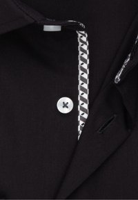 Camisa de tela negra con un botón blanco y detalle de ribete de patrón geométrico a lo largo del tapete. Incluye un bolsillo con muesca.