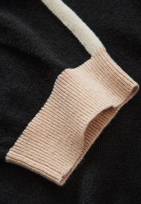 Dettaglio di un maglione lavorato a maglia con polsini a coste color pesca e corpo nero liscio a contrasto, che mette in risalto la variazione di texture e il contrasto di colore.