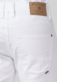 Los pantalones vaqueros de mezclilla blanca cuentan con una etiqueta de cuero marrón en la cintura, detalles en el bolsillo trasero y costuras en contraste. La tela tiene una textura suave.