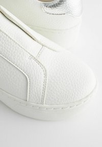 Białe sneakersy z teksturowaną skórzaną cholewką i błyszczącym srebrnym akcentem na pięcie. Okrągły nosek i masywna gumowa podeszwa.