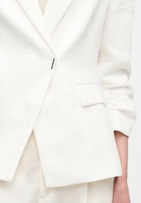 Λευκό, ραμμένο σακάκι με ένα μαύρο κουμπί, τσεπάκι με καπάκι στο πλευρό και ελαφρώς ανασηκωμένα μανίκια, φορεμένο πάνω από ταιριαστά παντελόνια.