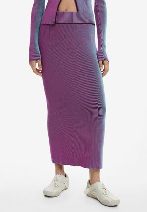 Femme portant une jupe midi côtelée à dégradé violet et bleu avec un haut à manches longues assorti et des baskets blanches à fermeture Velcro, debout sur un fond blanc.