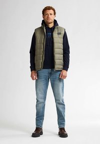 Khakifarbene Steppweste über einem marineblauen Hoodie, hellblaue Jeans und braune Schuhe. Verfügt über einen Reißverschluss und eine gesteppte Struktur.