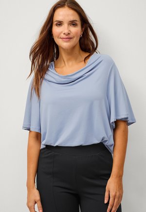 Light blue, short-sleeve top with a draped neckline, made from soft, smooth fabric. Paired with black, fitted pants.