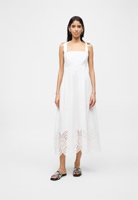 Γυναίκα που στέκεται φορώντας ένα λευκό μίντι φόρεμα χωρίς μανίκια με δεσίματα στους ώμους και τελείωμα από δαντέλα με μικρές οπές, συνδυασμένο με σανδάλια τύπου slides με σχέδιο φολιδών φιδιού.