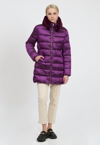 Frau trägt eine lange lila Daunenjacke mit Fellkragen, cremefarbene, cropped Hosen und schwarze Slipper mit goldener Kette, steht frontal.