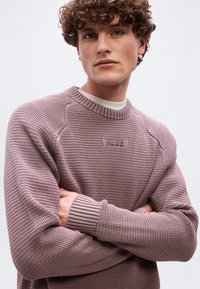 Νέος άνδρας με σγουρά μαλλιά, φοράει υφασμάτινο πουλόβερ σε αποχρώσεις του μοβ με το λογότυπο "HUGO", σταυρωμένα χέρια, ουδέτερη έκφραση σε ανοιχτόχρωμο φόντο.