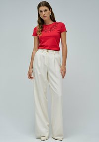 Camiseta roja de manga corta con adornos florales, combinada con pantalones blancos de talle alto y corte holgado, y tacones blancos de punta.