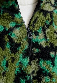 Fleece zippé vert et noir, avec un motif camouflage, présentant une fermeture éclair noire et un tissu texturé. Superposé à un vêtement de couleur claire.