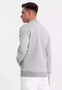 Sudadera gris claro con escote redondo y puños acanalados, que presenta un tejido texturizado y un corte relajado. Los bolsillos son visibles a los lados.