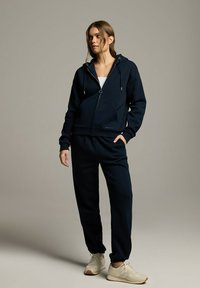 Granatowa bluza z kapturem na zamek błyskawiczny z troczkami, w połączeniu z dopasowanymi spodniami dresowymi. Oba elementy wykonane z miękkiego materiału w luźnym kroju i posiadające boczne kieszenie.