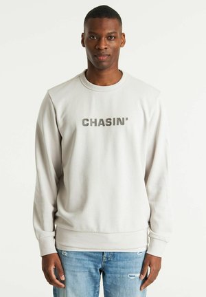 Felpa grigia chiara con polsini e orlo a coste. Presenta la scritta "CHASIN'" in grigio scuro sul petto. Indossata con jeans blu logori.