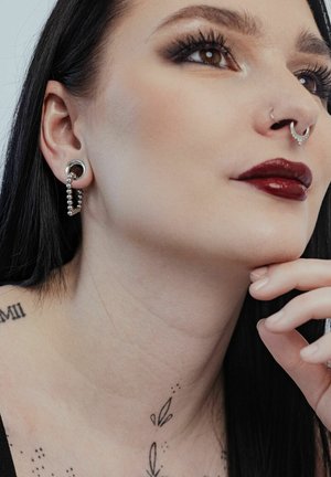 Silberner Ohrclip mit einer Kette aus kleinen Perlen. Nahaufnahme des Gesichts eines Models mit einem Septumring und dunklem Lippenstift. Glatte Hautstruktur.