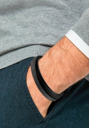 Black leather bracelet with smooth and textured bands, worn on a wrist. The hand is near a grey sweater and dark herringbone fabric.