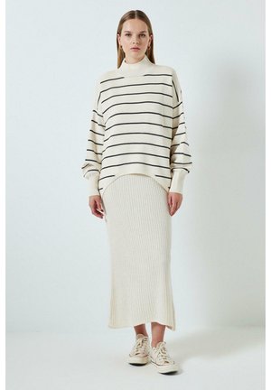 Nő fehér bordázott midi szoknyában, fehér, fekete vízszintes csíkokkal díszített bő pulóverben és fehér tornacipőben, egyszerű háttér előtt állva.