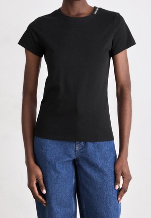 Musta, lyhythihainen vartalonmyötäinen t-paita, jossa pieni valkoinen logo kaula-aukon lähellä. Yhdistettysinisiin farkkuihin, seisovan henkilön päällä yksinkertaista taustaa vasten.