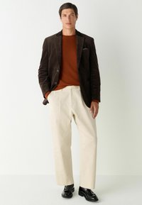 Een corduroy bruine blazer over een oranje gebreid sweater, gecombineerd met crèmekleurige broek en zwarte schoenen. Opvallende details zijn de knopen en zakken van de blazer.