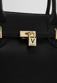 Svart texturerad handväska med ett guldfärgat lås med nyckelhål, en logotavla och dubbla handtag för bärande.