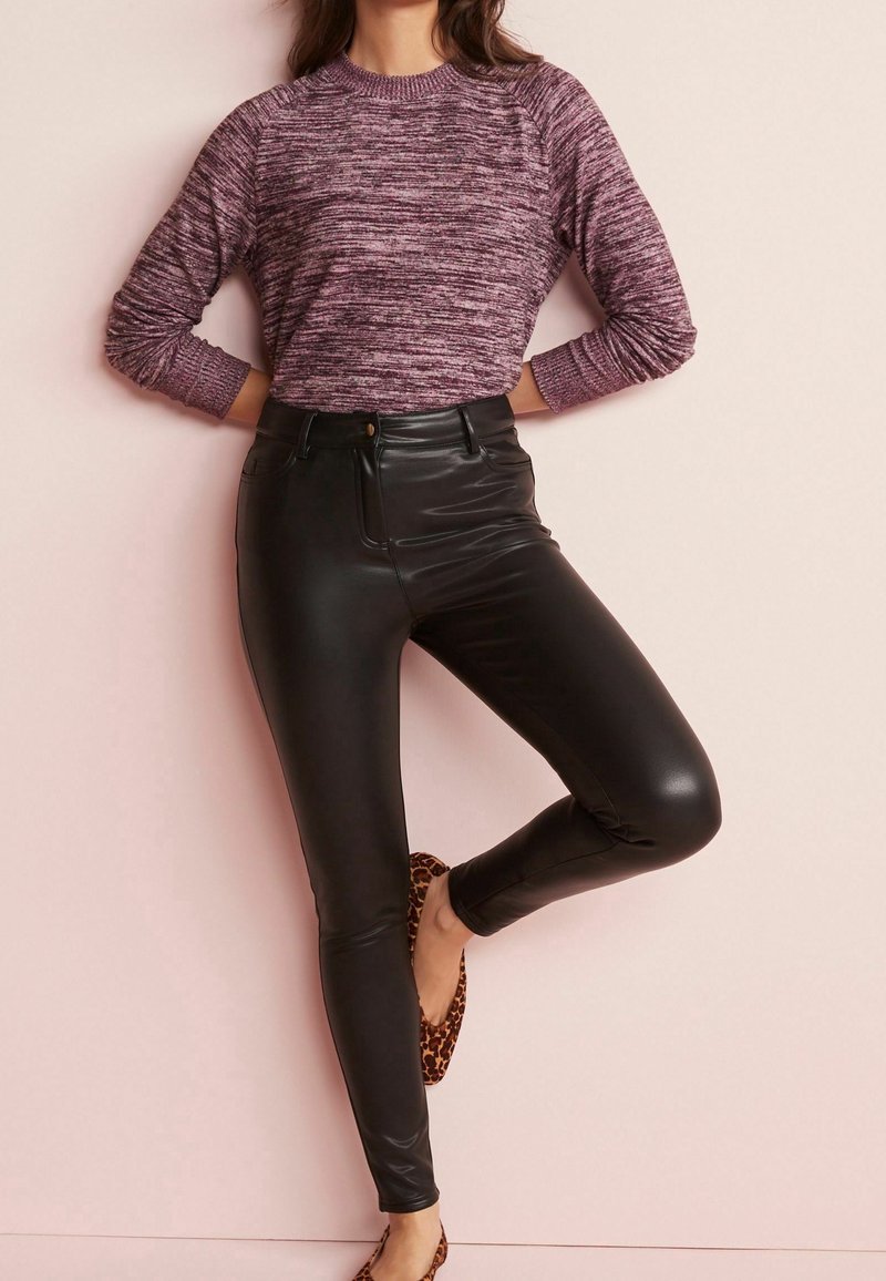 Femme portant un pull violet chiné, un pantalon noir taille haute en similicuir et des ballerines à imprimé léopard, debout contre un mur beige.