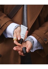 Tube blanc écrasable de gel pour les mains étiqueté "MARIETTE LALA MARIE Lemon Notes" tenu en main, avec un bouchon noir en train d'être retiré. Manteau marron et pull gris visibles.