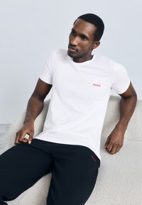 Baltas medvilninis marškinėlis su raudonu „HUGO“ logotipu kairėje krūtinės pusėje, derinamas su juodomis tekstūruotomis kelnėmis. Sėdi ant šviesios spalvos, išlenktos kėdės.