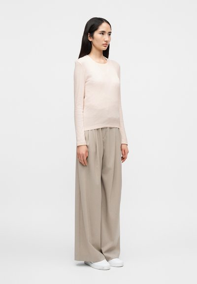 Γυναίκα που στέκεται, φοράει μακρυμάνικο μπλουζάκι σε ανοιχτό ροζ χρώμα, φαρδιά μπεζ παντελόνια και λευκά παπούτσια χωρίς κορδόνια, μπροστά σε ένα απλό γκρι φόντο.