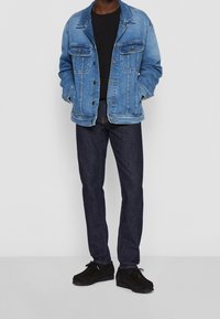 Chaqueta vaquera azul con dos bolsillos en el pecho, camiseta negra debajo, vaqueros oscuros y zapatos negros. Diseño y colores simples y clásicos.