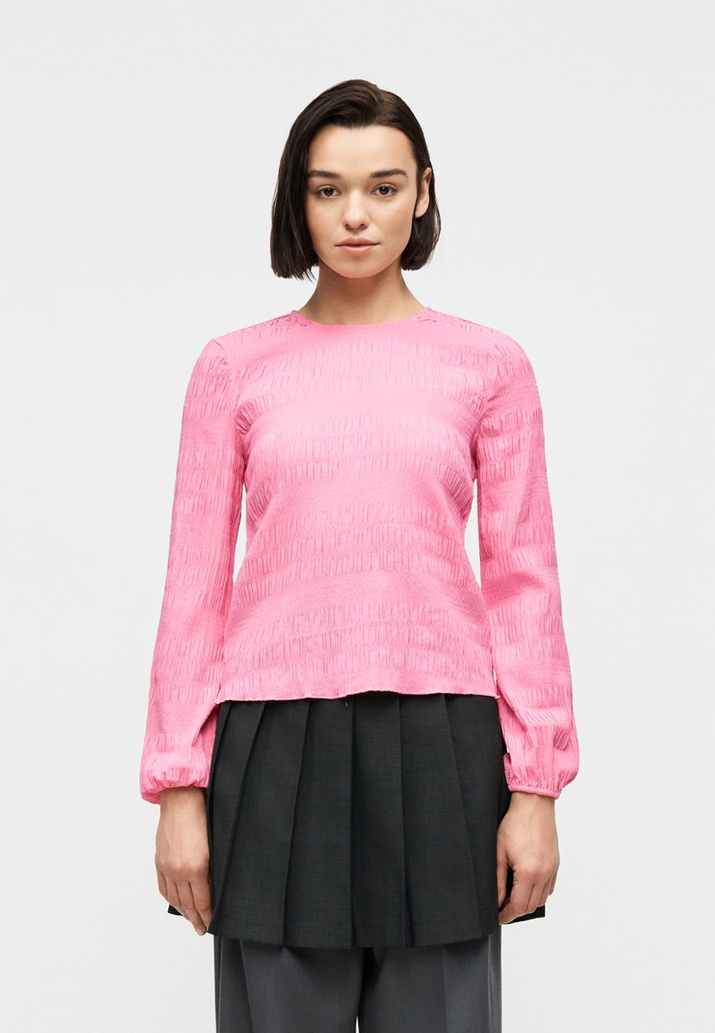 Γυναίκα με κοντά σκούρα μαλλιά που φοράει ροζ μπλούζα με υφή και μακριά μανίκια και μαύρη πλισέ φούστα πάνω από φαρδιά γκρι παντελόνια, στραμμένη προς τα εμπρός.