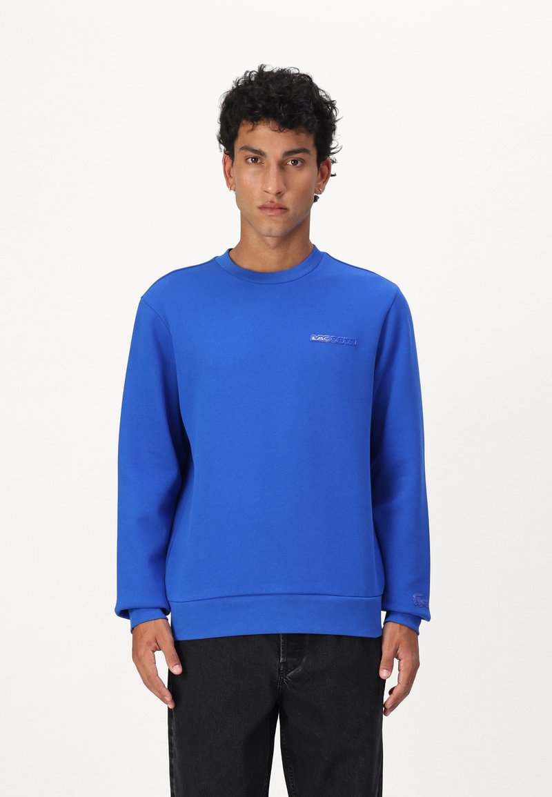 Sudadera azul brillante con cuello redondo y mangas largas. Presenta un pequeño logo en un color contrastante en el pecho. Textura suave.