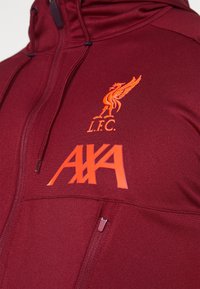 Bordeauxfarbener Hoodie mit Reißverschluss, versehen mit einem orangefarbenen, gestickten Liverpool FC-Logo und dem Text "AXA" auf der Vorderseite. Glatte Stoffstruktur ohne Muster.