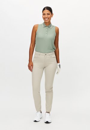 Uśmiechnięta kobieta w bezrękawniku do golfa w kolorze szałwi zielonej, beżowych spodniach, białej rękawicy golfowej na lewej ręce oraz białych trampkach stoi na białym tle.