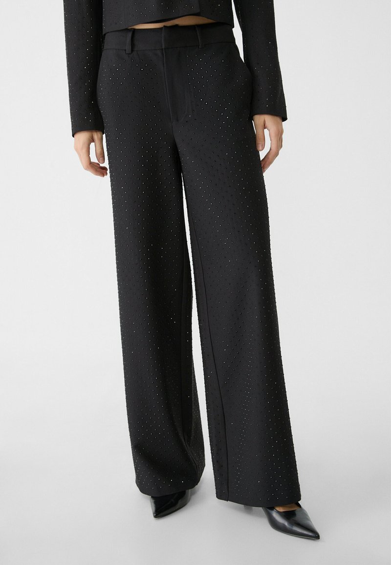 Pantalones de pierna ancha negros con una superficie texturizada que presenta embellishments en forma de puntos. Completos con una cinturilla ajustada y pliegues en la parte delantera.
